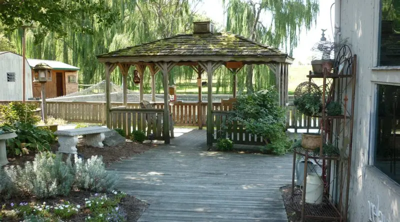 gazebo en bois avec toit recouvert de mousse, entouré de saules pleureurs et d'un patio en bois