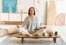 Woman peacefully meditating indoors surrounded by cushions and tea set, promoting tranquility.