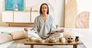 Woman peacefully meditating indoors surrounded by cushions and tea set, promoting tranquility.