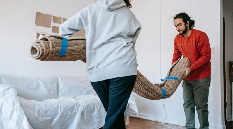 A couple unrolls a carpet in their new apartment, amidst moving and unpacking.